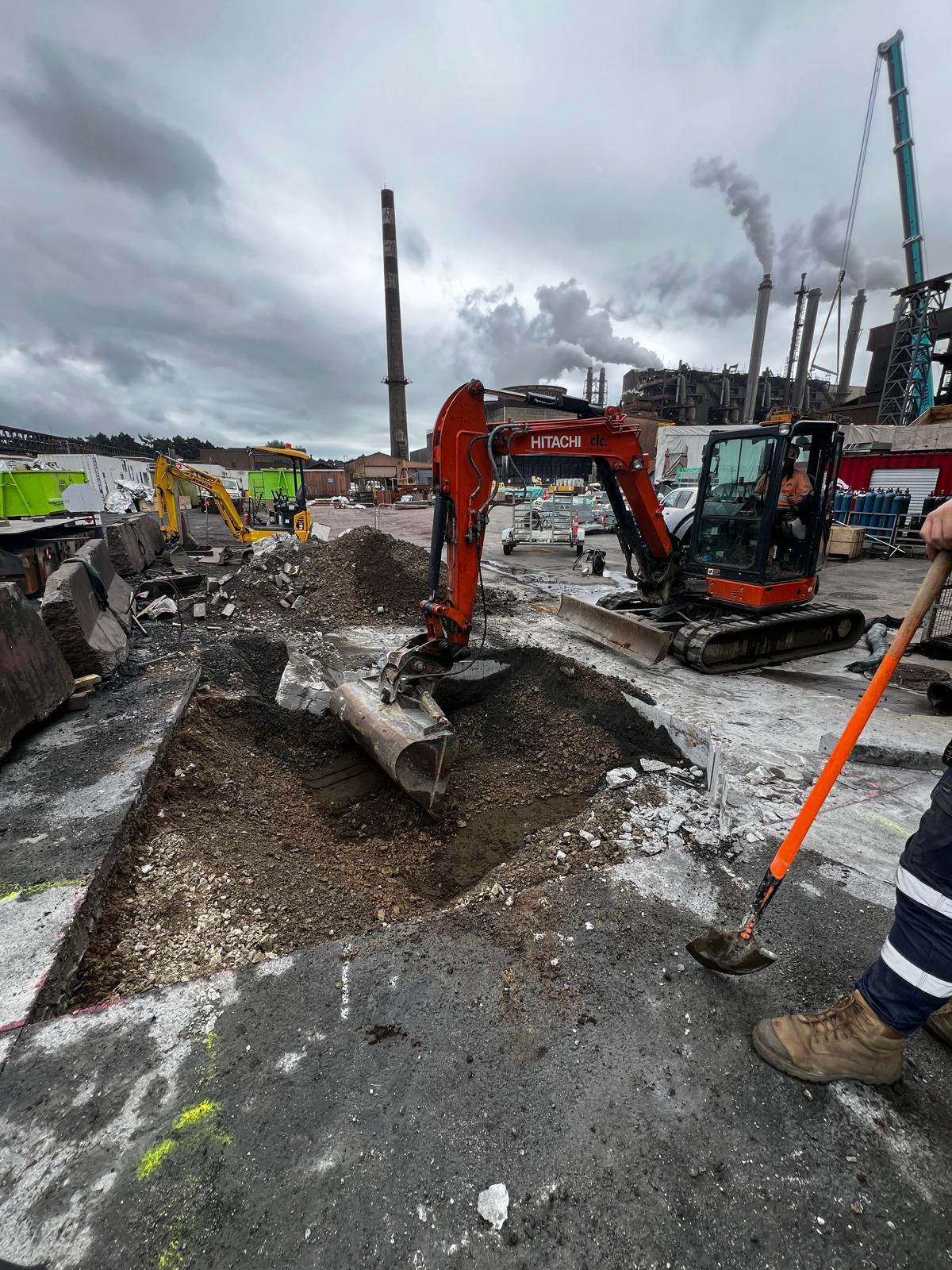 Excavator working beside an open excavation and exposed service trench on an urban construction site