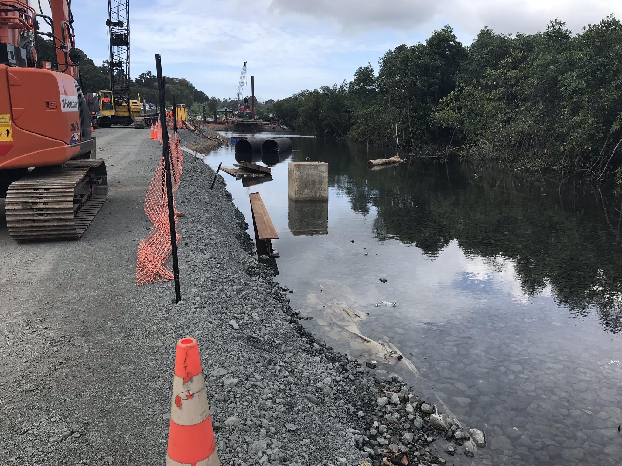 Crawler crane and excavator working on a gravel platform beside a tidal waterway, with a piling barge in the background